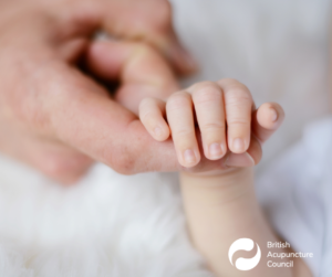 A small baby hand lays on a white fluffy blanket whilst reaching hold to hold it's mothers index finger.